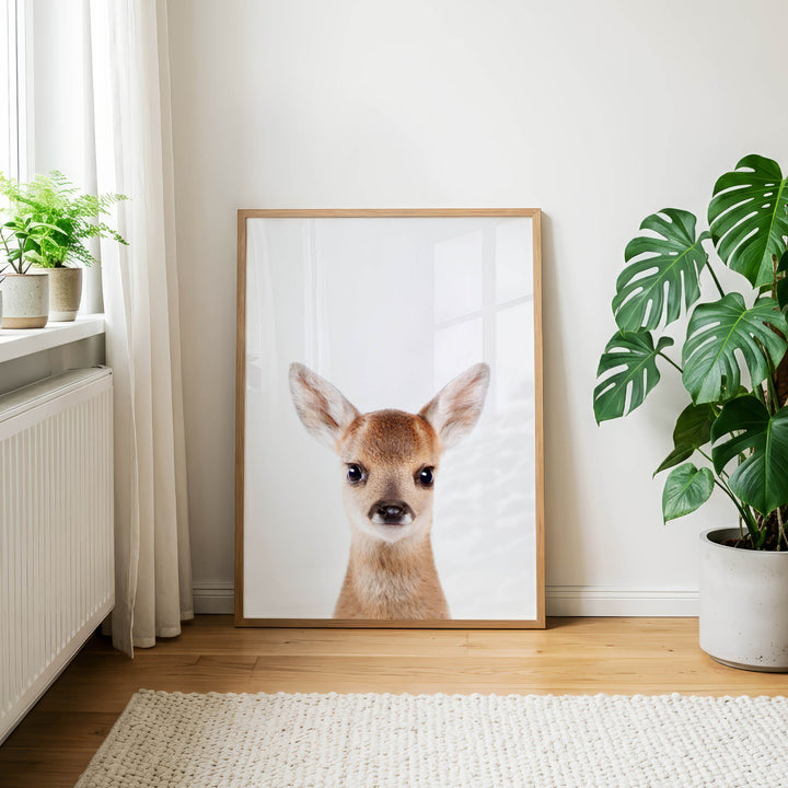 a large framed picture of a deer with a white background and a wooden frame, placed on a wooden floor in a room with a window and potted plants.