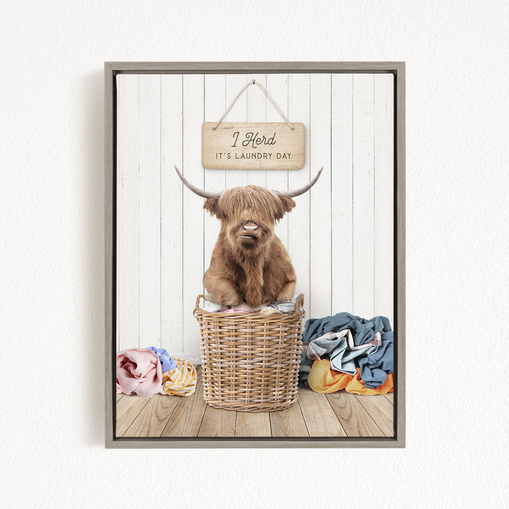 A Highland cow sits in a wicker basket, surrounded by laundry, with a sign above that reads "I herd it's laundry day".