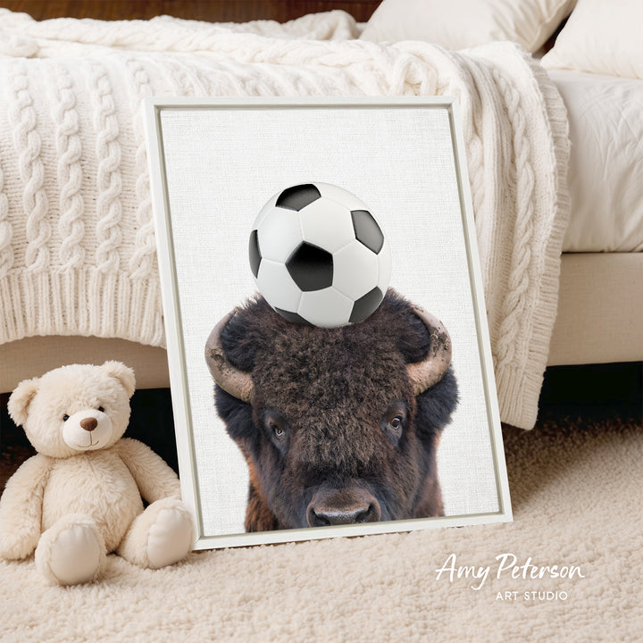 A framed image of a buffalo with a soccer ball on its head, placed on a bed next to a stuffed teddy bear.