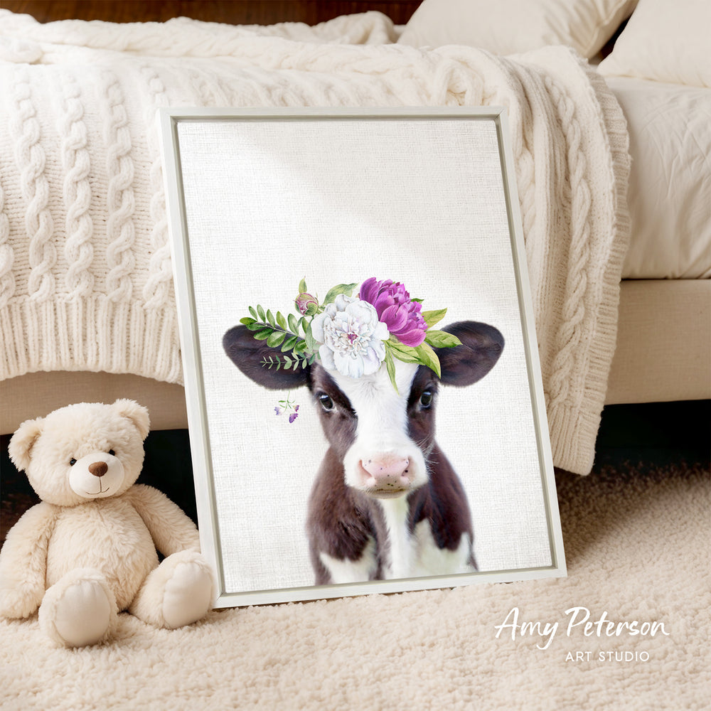 A framed print of a cow wearing a flower crown is displayed on a bed, accompanied by a teddy bear.