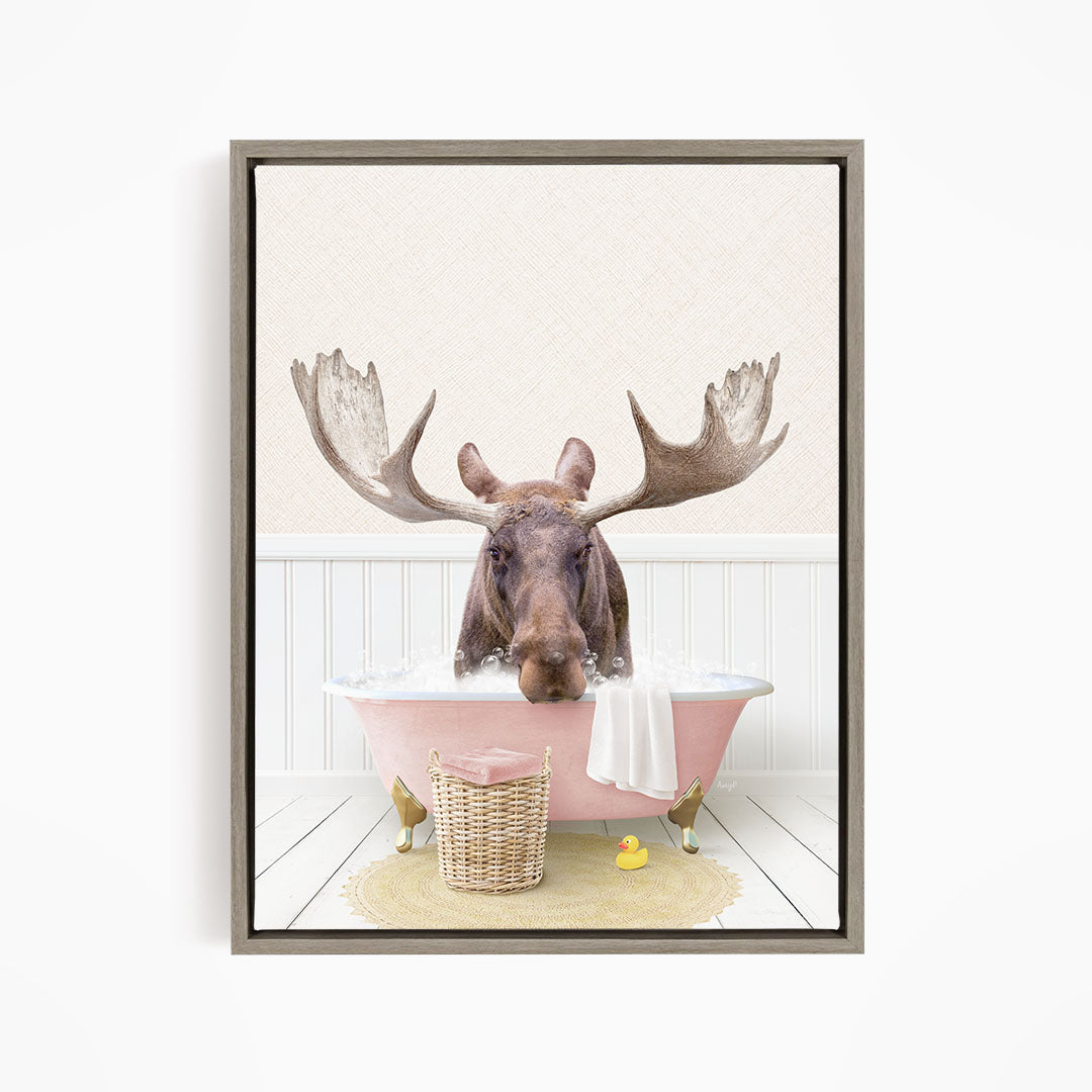 A moose with large antlers is sitting in a pink bathtub filled with water, surrounded by a yellow rubber duck and a basket.