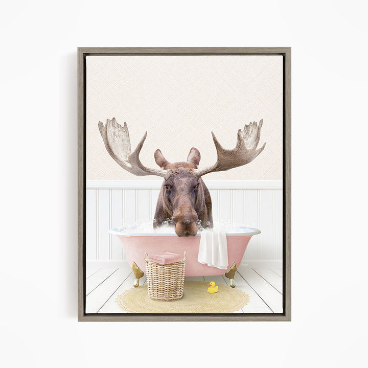 A moose with large antlers is sitting in a pink bathtub filled with water, surrounded by a yellow rubber duck and a basket.