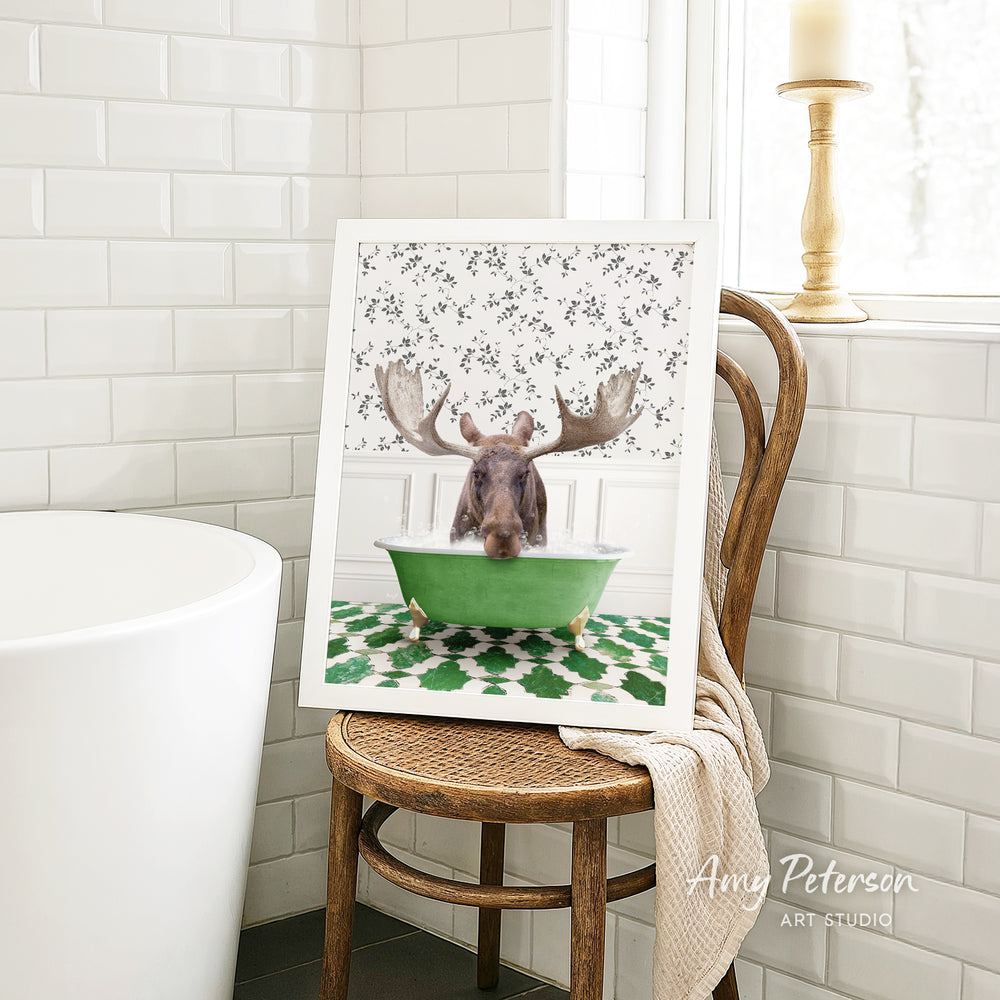 A framed image of a moose sitting in a green bathtub, with a floral pattern on the walls behind it, is displayed on a wooden chair in a bathroom setting.