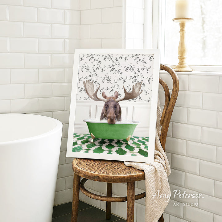 A framed image of a moose sitting in a green bathtub, with a floral pattern on the walls behind it, is displayed on a wooden chair in a bathroom setting.