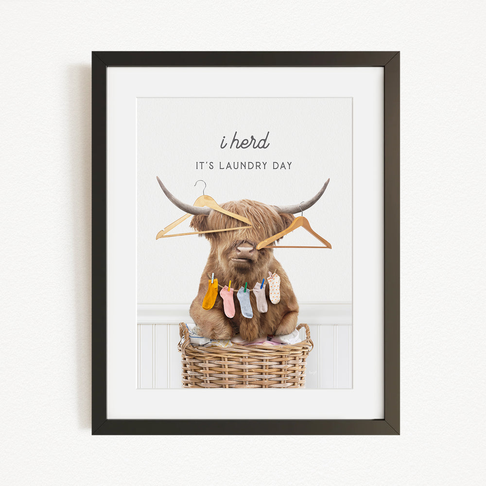 A framed image depicting a Highland cow sitting in a wicker basket, surrounded by clothes and hangers, with the text "It's laundry day" above it.