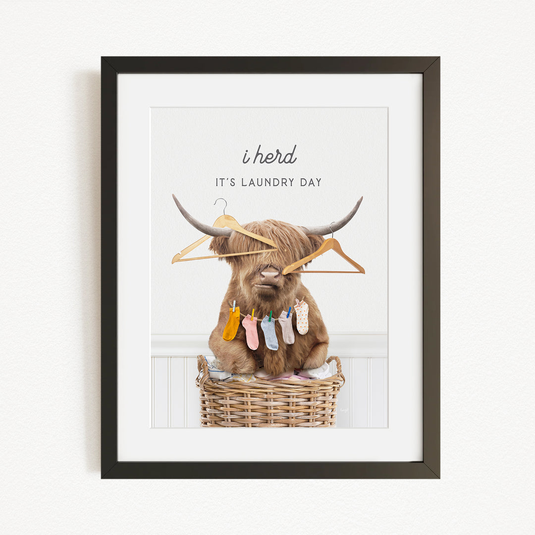 A framed image depicting a Highland cow sitting in a wicker basket, surrounded by clothes and hangers, with the text "It's laundry day" above it.