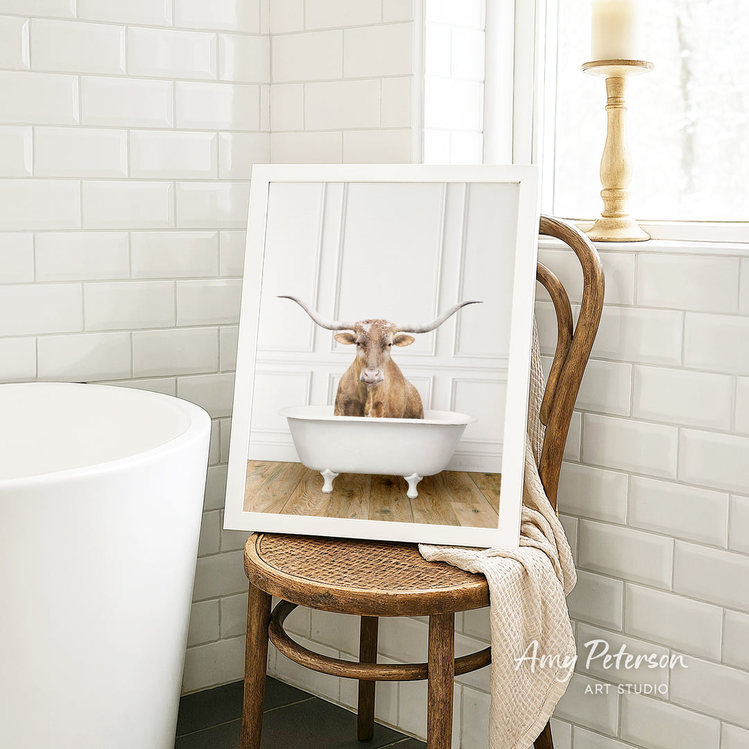 A framed image of a cow sitting in a bathtub is displayed on a wooden chair in a bathroom setting.