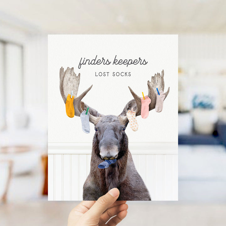 A hand holding a card with a moose wearing socks and the text "finders keepers" and "lost socks" printed on it.