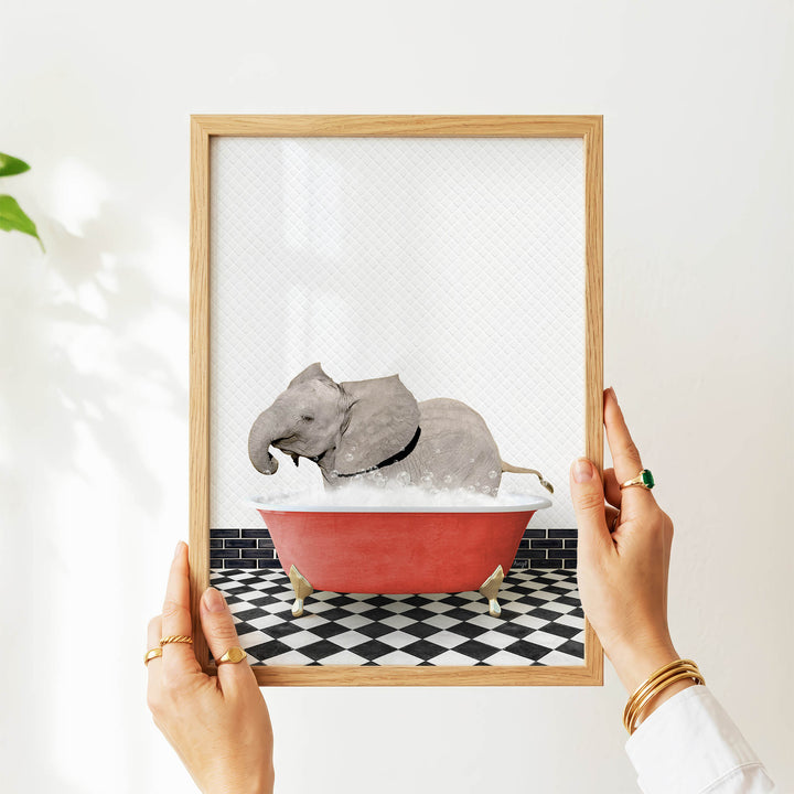 A person's hands are holding a framed picture of a gray elephant sitting in a red bathtub on a black and white checkered floor.