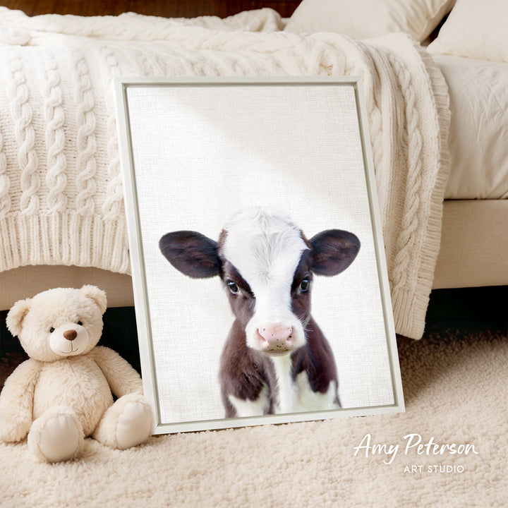 A framed image of a black and white cow is displayed on a white wall, accompanied by a teddy bear on a beige carpet.