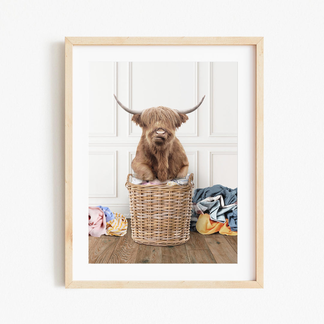 A Highland cow with long horns is sitting in a wicker basket on a wooden floor, surrounded by clothes.