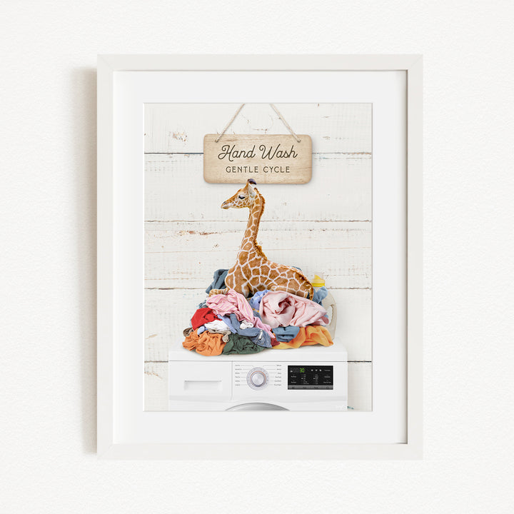 A framed image depicting a giraffe sitting atop a pile of laundry, with a sign above that reads "Hand Wash Gentle Cycle".
