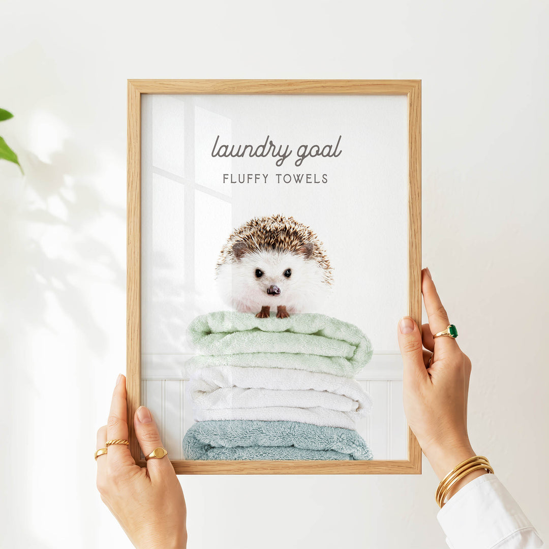A person is holding up a framed picture of a hedgehog sitting on a stack of fluffy towels, with the text "laundry goal" and "fluffy towels" written above the image.