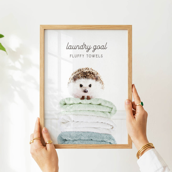 A person is holding up a framed picture of a hedgehog sitting on a stack of fluffy towels, with the text "laundry goal" and "fluffy towels" written above the image.
