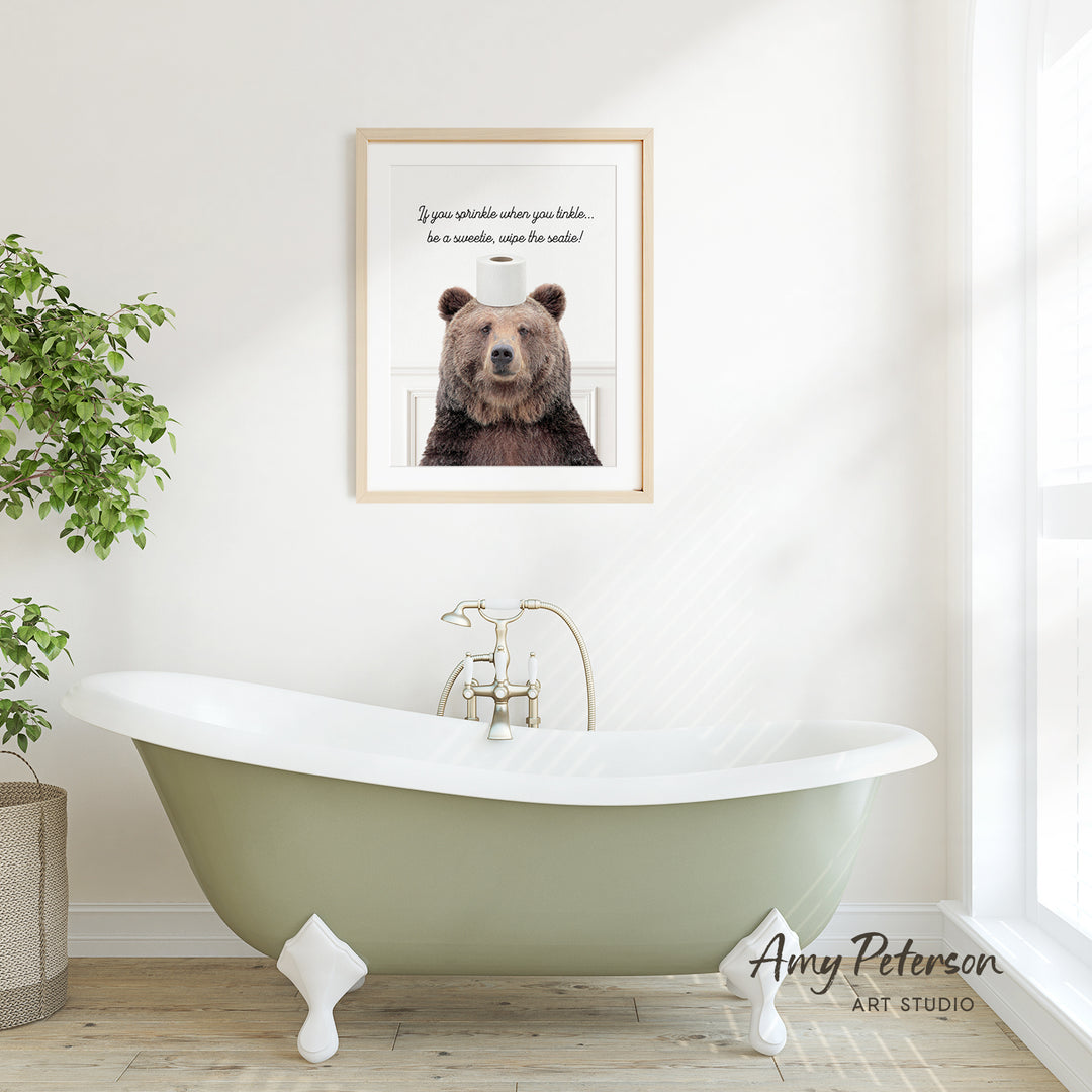 a framed picture of a brown bear with a humorous caption above it, which reads "If you spill water when you sneeze, be sure to wipe the nose!" The picture is displayed above a green clawfoot bathtub in a bathroom setting.