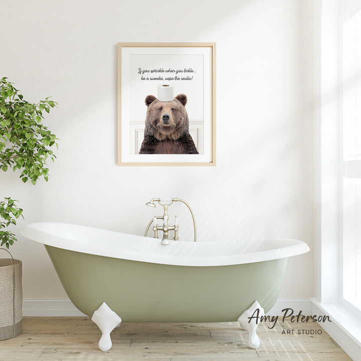 a framed picture of a brown bear with a humorous caption above it, which reads "If you spill water when you sneeze, be sure to wipe the nose!" The picture is displayed above a green clawfoot bathtub in a bathroom setting.