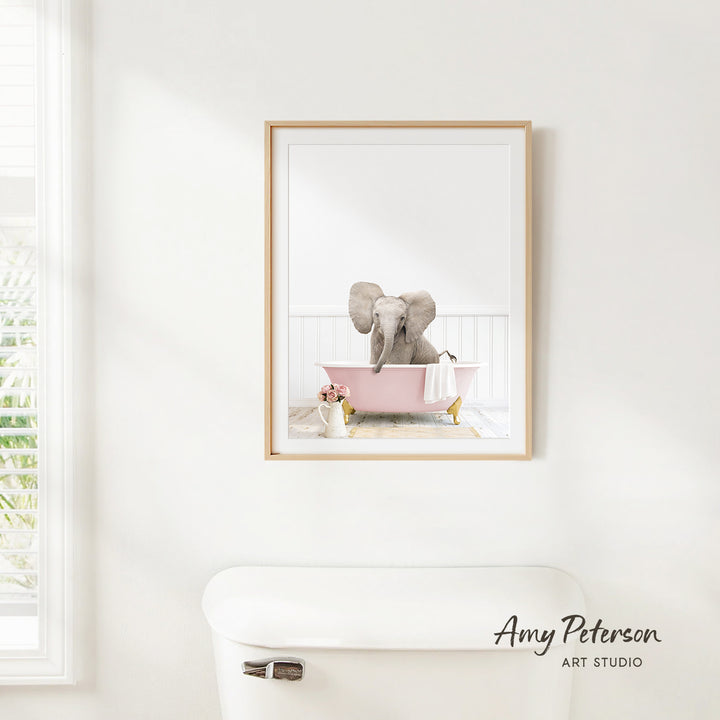 A framed image of an elephant sitting in a pink bathtub, with a window and toilet visible in the background.