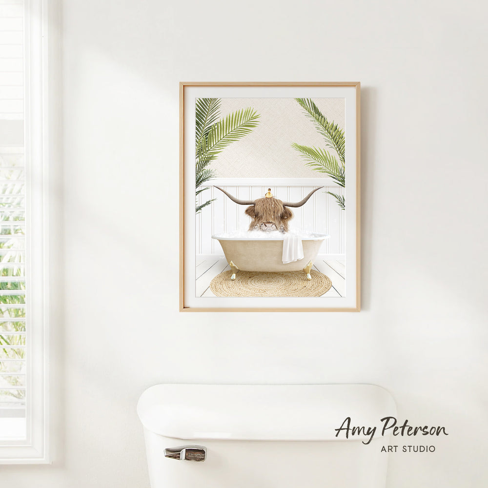 A framed print of a cow sitting in a bathtub, surrounded by palm leaves, is hanging on a white wall in a bathroom.