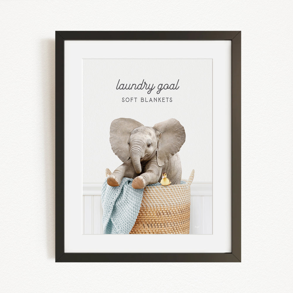 A framed image of a baby elephant sitting in a woven basket, with a blanket and a yellow duck nearby.