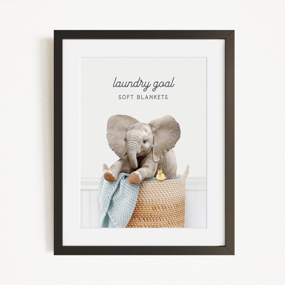 A framed image of a baby elephant sitting in a woven basket, with a blanket and a yellow duck nearby.