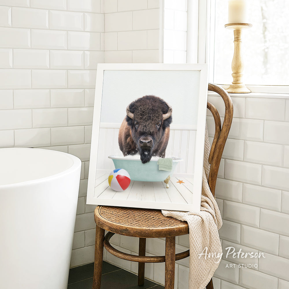 A framed image of a bison standing in a bathtub, surrounded by a beach ball, a starfish, and a towel, is displayed on a wooden chair in a bathroom setting.