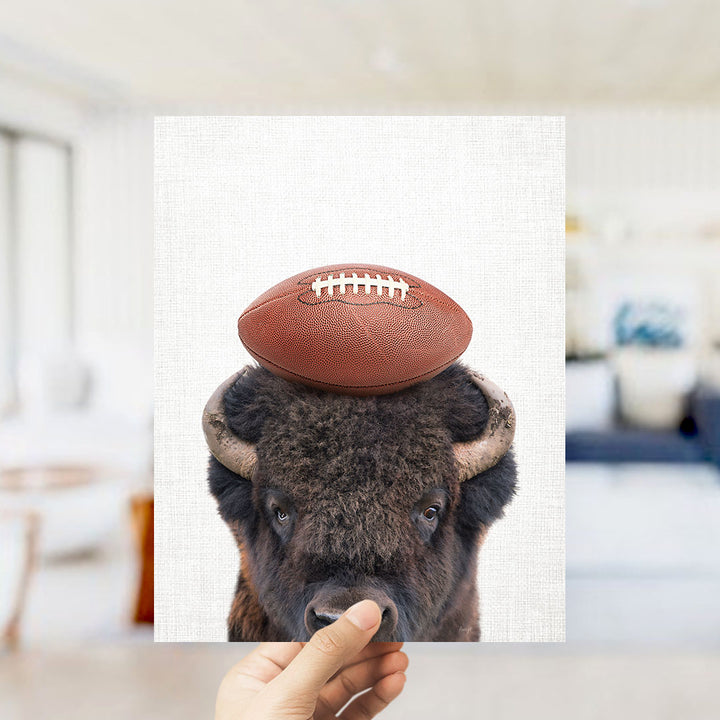 A hand holds up a framed image of a buffalo with a football on its head.