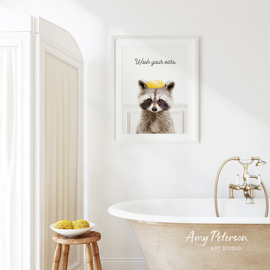 a bathroom with a framed picture of a raccoon wearing a yellow hat, a wooden stool with a bowl of fruit, and a bathtub with a faucet.