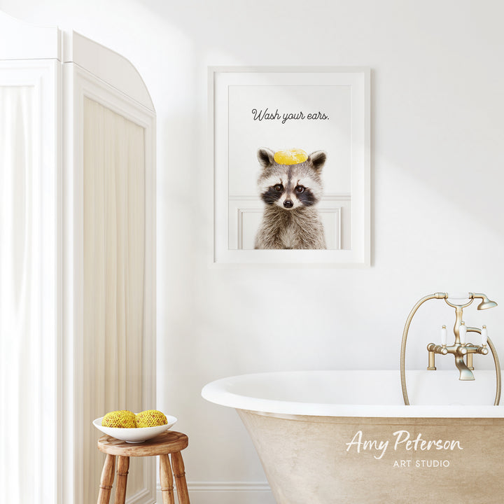 a bathroom with a framed picture of a raccoon wearing a yellow hat, a wooden stool with a bowl of fruit, and a bathtub with a faucet.