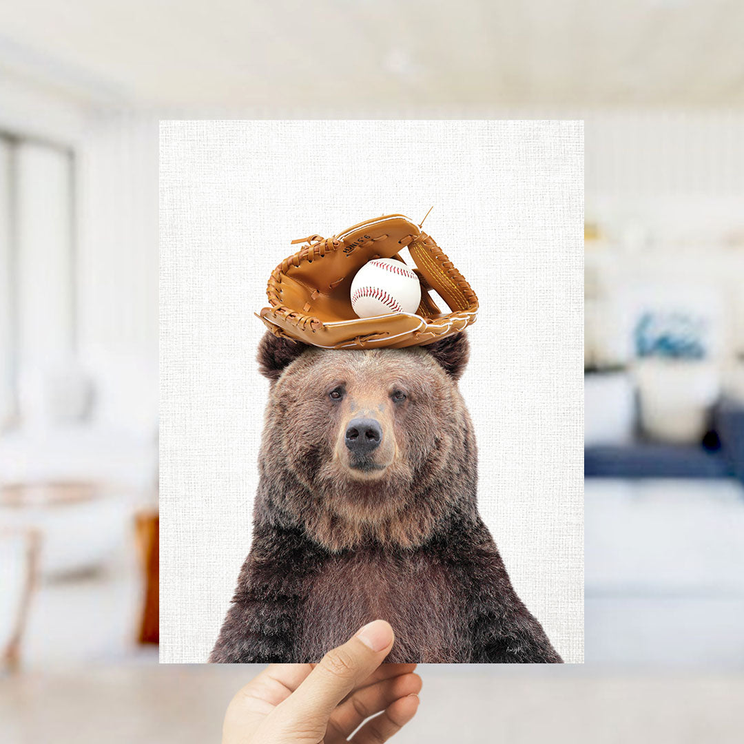 A hand holds up a framed image of a brown bear wearing a baseball glove and holding a baseball on its head.