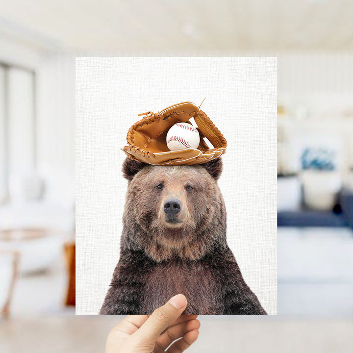 A hand holds up a framed image of a brown bear wearing a baseball glove and holding a baseball on its head.