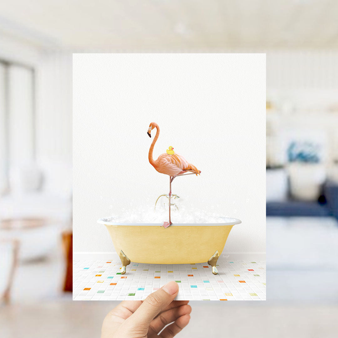 A hand holds up a framed image of a pink flamingo standing on a yellow bathtub filled with bubbles, with a blurred background suggesting a bathroom setting.