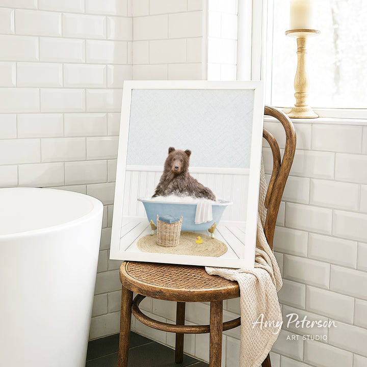 A framed image of a brown bear sitting in a blue bathtub, surrounded by a basket of bath toys, is displayed on a wooden chair in a bathroom setting.