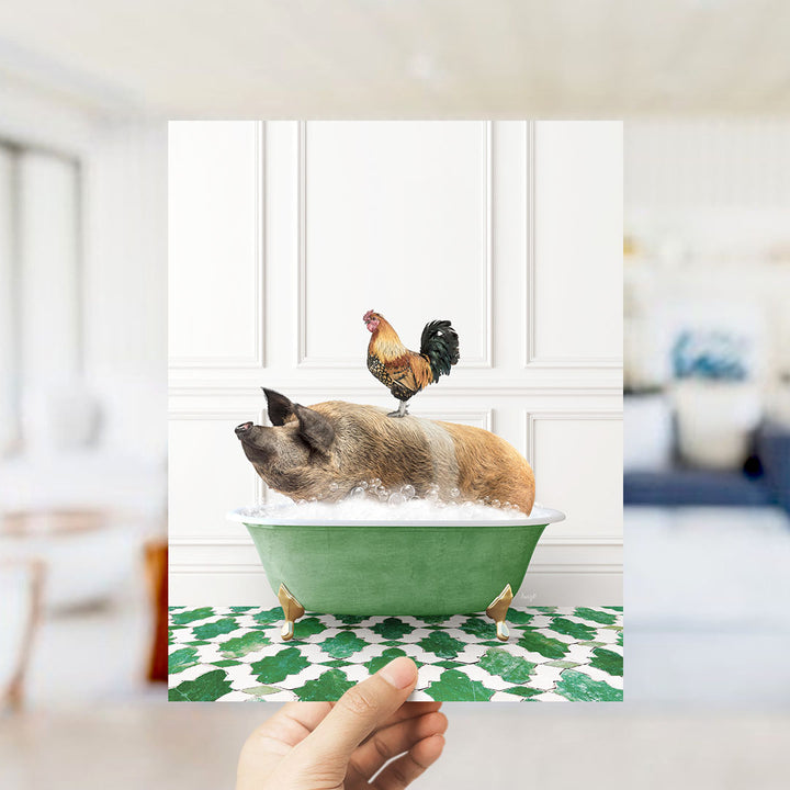 A hand holds up a framed image depicting a pig sitting in a green bathtub, with a rooster perched on its back.