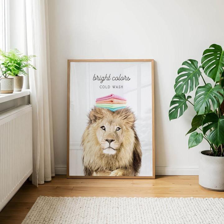 a framed poster of a lion with a stack of colorful towels on its head, placed on a wooden floor in a room with a window and potted plants.