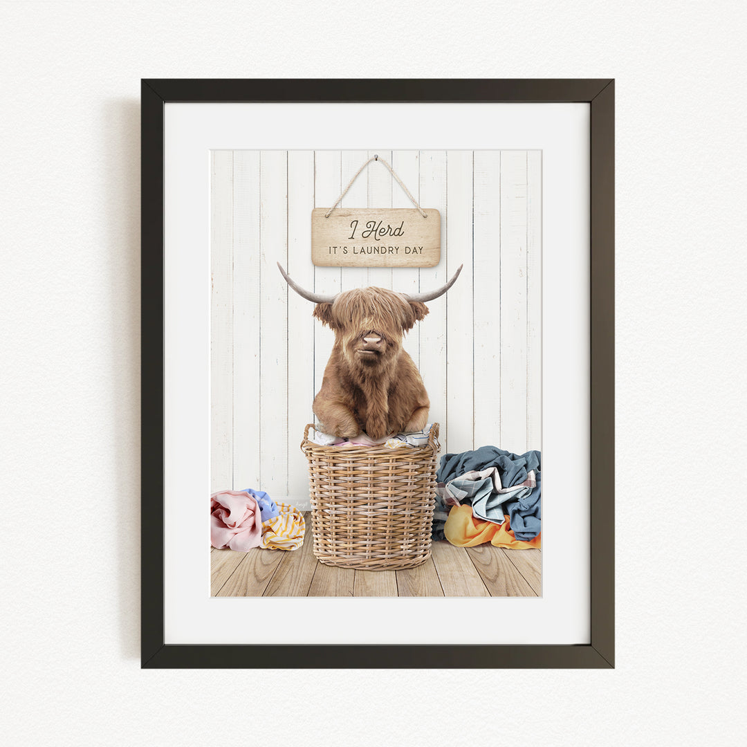A framed image of a Highland cow sitting in a wicker basket, with a sign above it that says "It's laundry day".