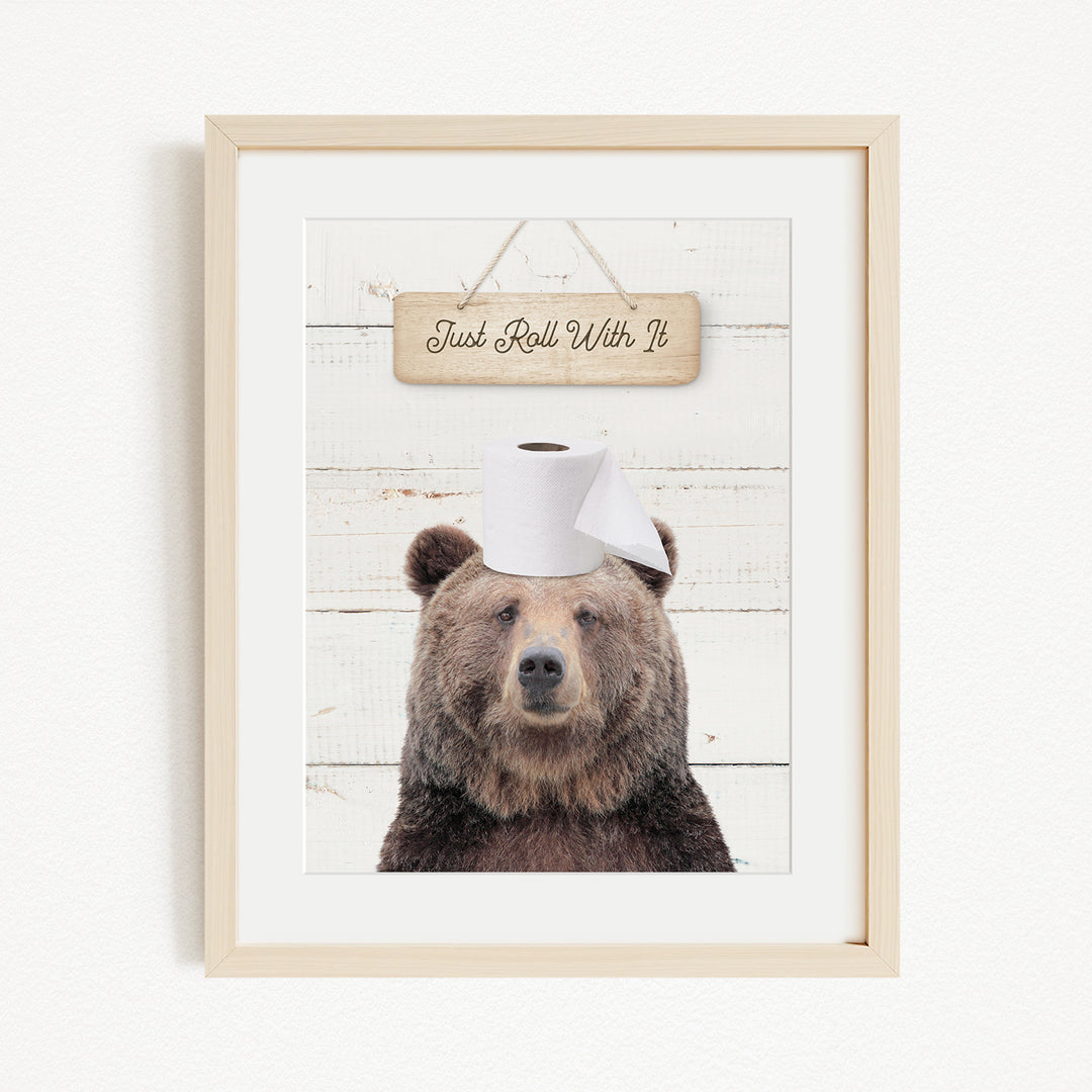 A framed image of a brown bear wearing a white toilet paper hat, with a wooden sign above it that says "Just Roll With It".