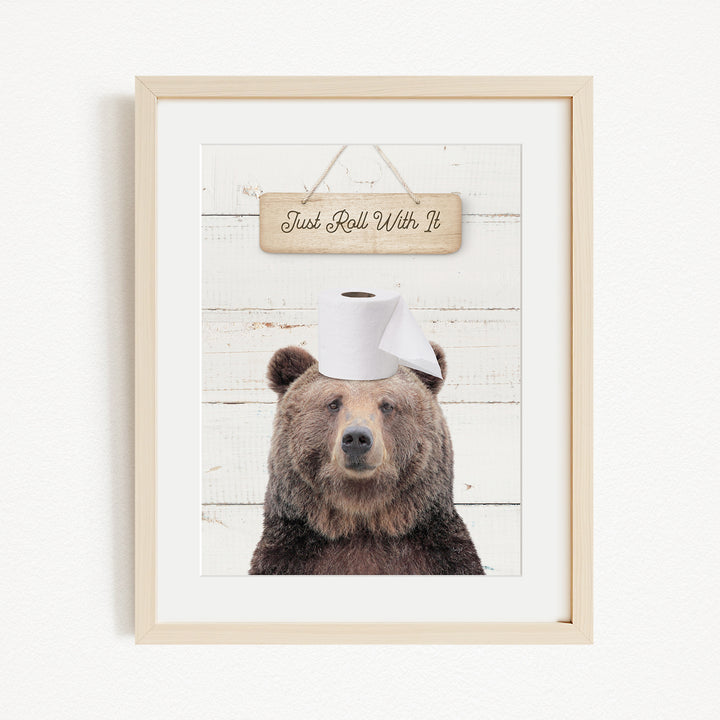 A framed image of a brown bear wearing a white toilet paper hat, with a wooden sign above it that says "Just Roll With It".