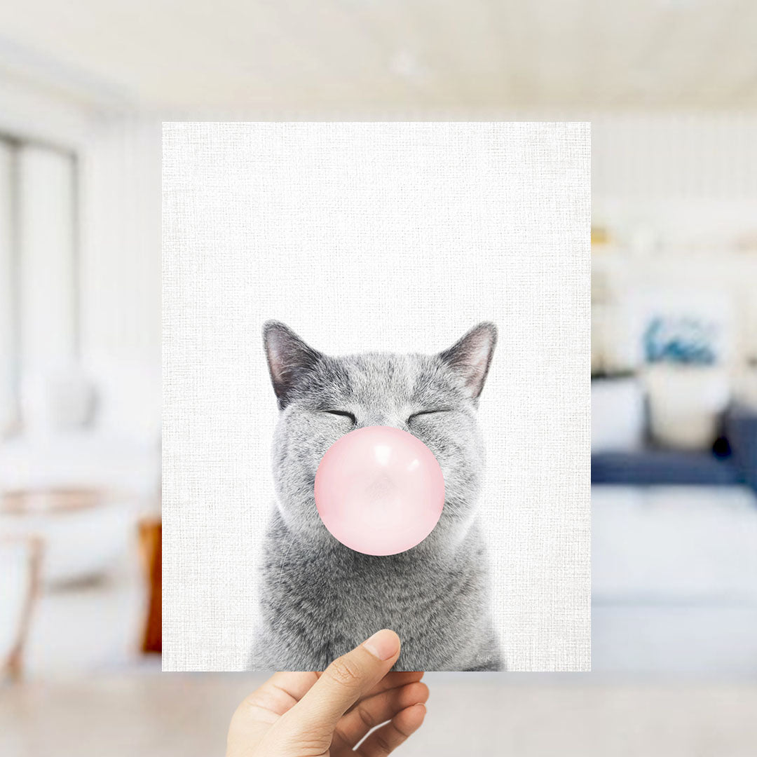 A gray cat is blowing a pink bubble with its mouth, as a person holds up a framed print of the image.