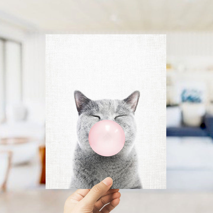 A gray cat is blowing a pink bubble with its mouth, as a person holds up a framed print of the image.