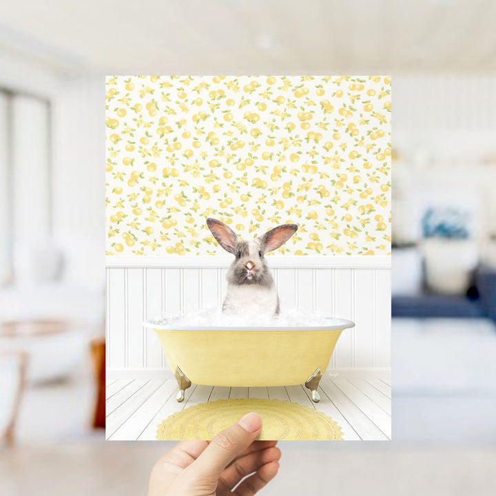 A hand holds up a photograph of a rabbit sitting in a yellow bathtub filled with bubbles, surrounded by a yellow and white floral pattern on the wall.