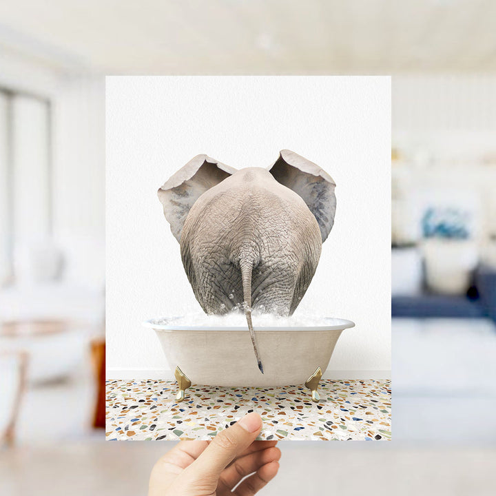 A hand holds up a framed image of an elephant standing in a bathtub filled with water, creating a whimsical and playful scene.