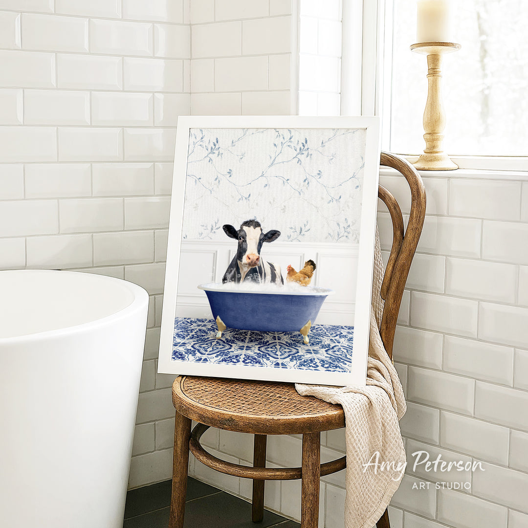 A framed image of a cow and a chicken sitting in a blue bathtub, with a towel draped over the back of a wooden chair in a bathroom setting.