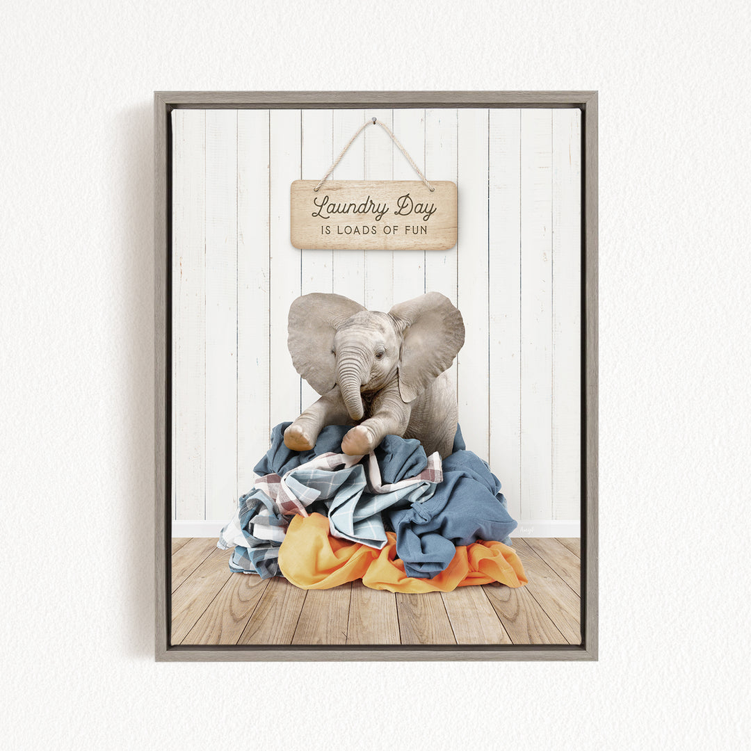 A framed image of a stuffed elephant sitting on a pile of laundry, with a wooden sign above it that says "Laundry Day is loads of fun".