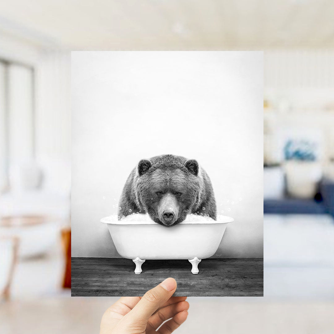 A hand holds up a black and white photograph of a bear sitting in a bathtub, with a blurred background suggesting a bathroom setting.