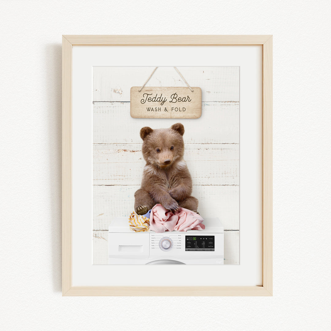 A framed image of a teddy bear sitting on a white washing machine, with a sign above it that says "Teddy Bear Wash & Fold".