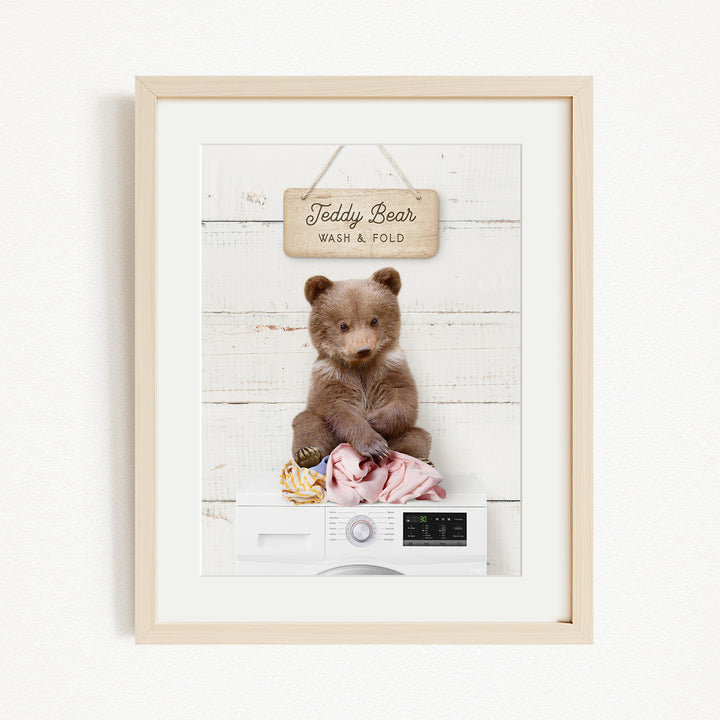 A framed image of a teddy bear sitting on a white washing machine, with a sign above it that says "Teddy Bear Wash & Fold".