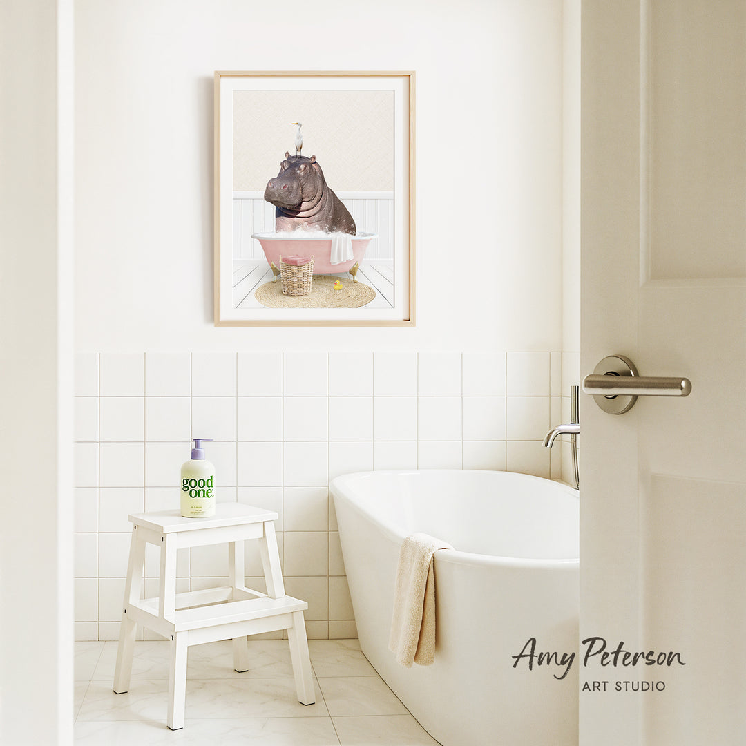 A framed illustration of a hippopotamus sitting in a bathtub is displayed on the wall above a white bathtub in a bathroom.
