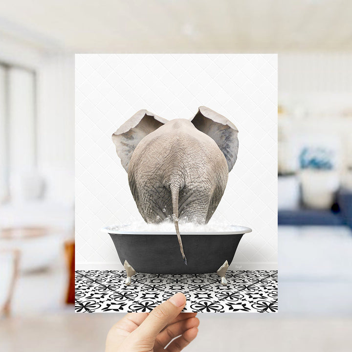 A hand holds up a framed image of an elephant sitting in a bathtub filled with water, creating a whimsical and playful scene.