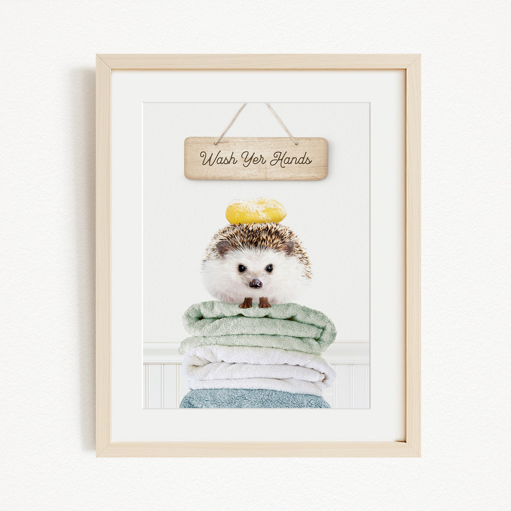 A framed image of a hedgehog sitting atop a stack of folded towels, with a wooden sign above that reads "Wash Your Hands".