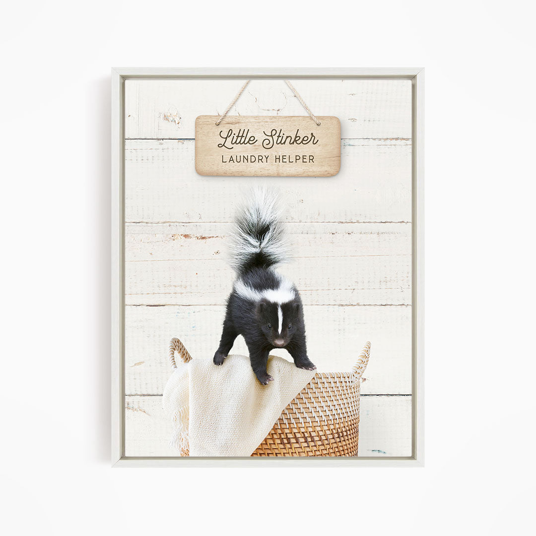 A black and white skunk standing on a woven basket, with a wooden sign above that reads "Little Stinker Laundry Helper".